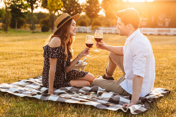 Image of young loving couple sitting by dating outdors on picnic holding glasses of wine drinking. - Φωτογραφία, εικόνα