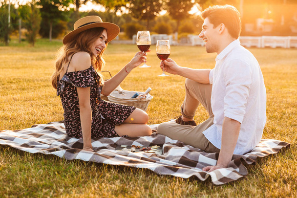 Image of young loving couple sitting by dating outdors on picnic holding glasses of wine drinking. - Foto, imagen