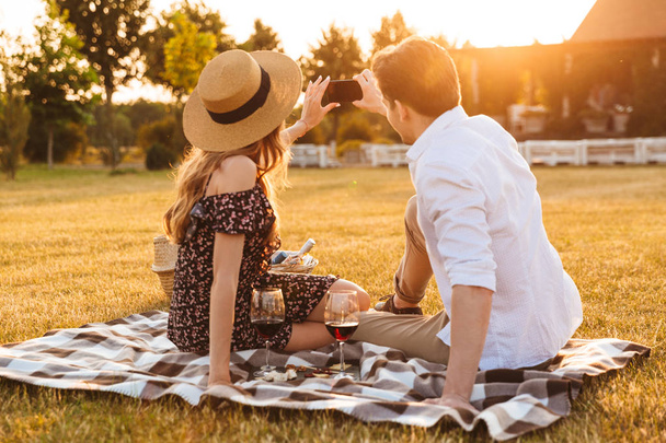 Photo of young cute loving couple sitting by dating outdors on picnic holding glasses of wine take a selfie by mobile phone. - Fotografie, Obrázek