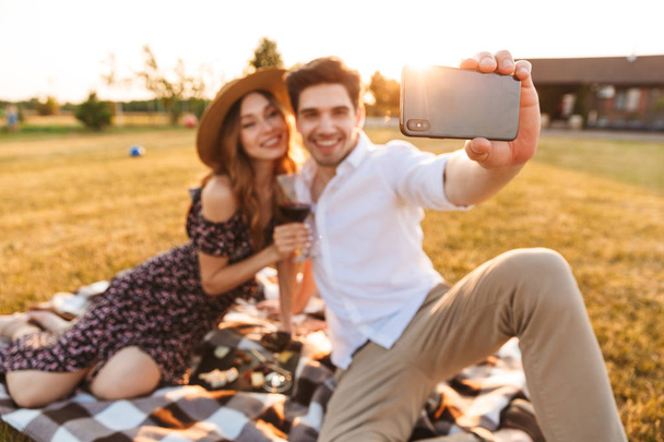 Photo of young cute loving couple sitting by dating outdors on picnic holding glasses of wine take a selfie by mobile phone. - Fotó, kép