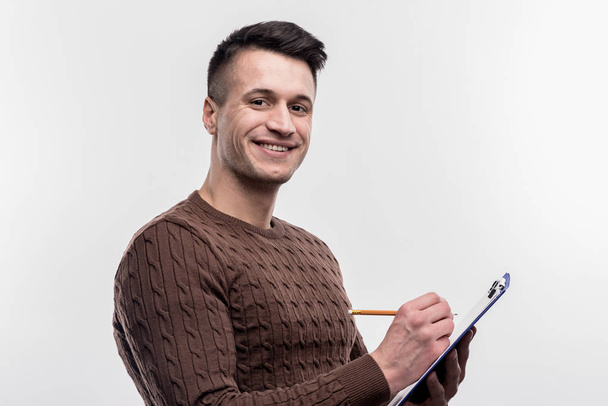Χαμογελώντας μελαχρινός άνδρες Γραμματέας φοράει καφέ πουλόβερ κάνοντας σημειώσεις - Φωτογραφία, εικόνα