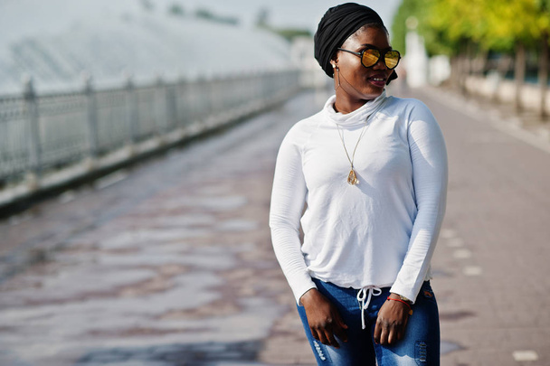 African muslim girl in sunglasses, black hijab, white sweatshirt and jeans posed outdoor against fountain. - 写真・画像