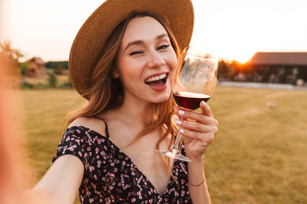 Image of cute pretty young woman outdoors holding glass drinking wine make a selfie by camera winking. - Foto, Imagen