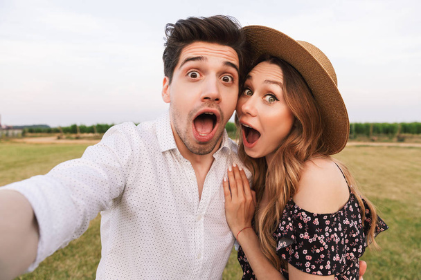 Joyful beautiful couple man and woman dating and taking selfie together while walking outdoor through field - Fotó, kép