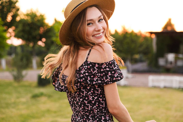 Image of cheerful cute woman wearing straw hat and dress smiling while walking outdoor on nature in countryside - Foto, afbeelding