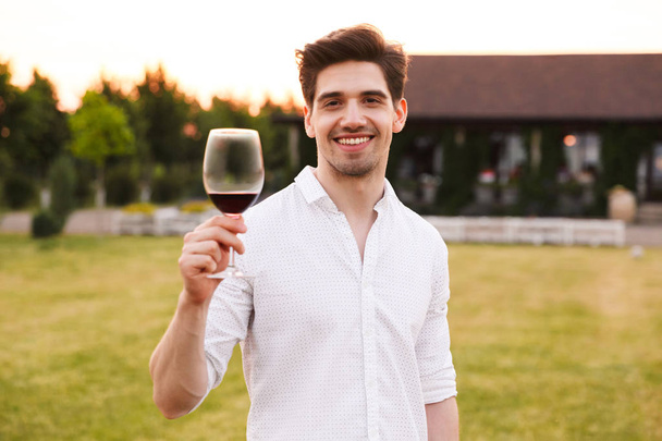 Photo of cheerful young guy wearing white shirt smiling and holding glass of wine while walking outdoor in countryside - Zdjęcie, obraz