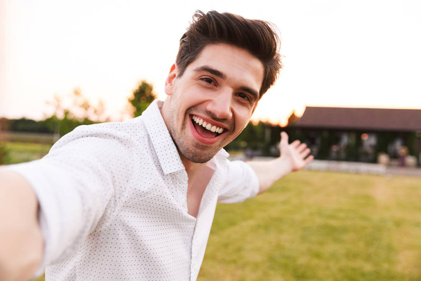Image of happy adult man wearing white shirt laughing while taking selfie during walk on nature near country house - Foto, immagini
