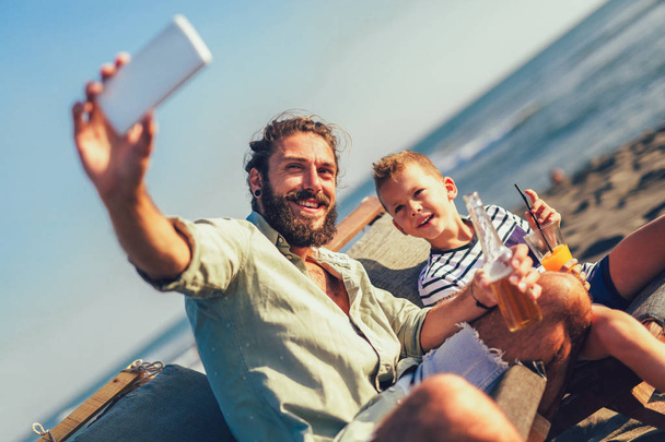 Aufnahme von Vater und Sohn beim Selfie am Strand. Junger Mann mit kleinem Jungen macht Selbstporträt mit Smartphone am Meer. - Foto, Bild