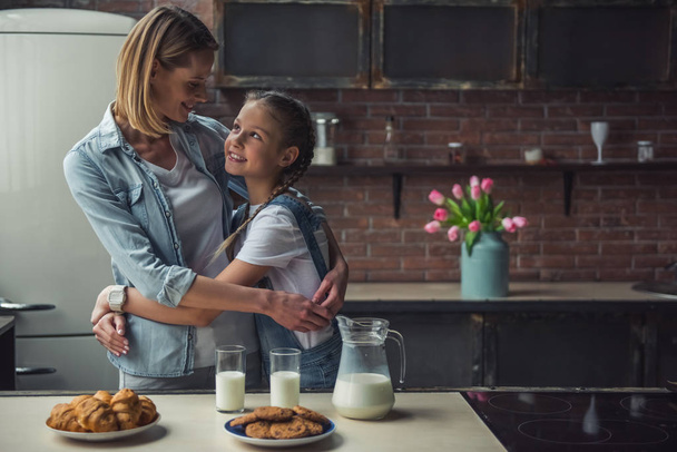 Mom and daughter in casual clothes are looking at each other and smiling while hugging in kitchen at home; cookies and milk on the table - Fotografie, Obrázek