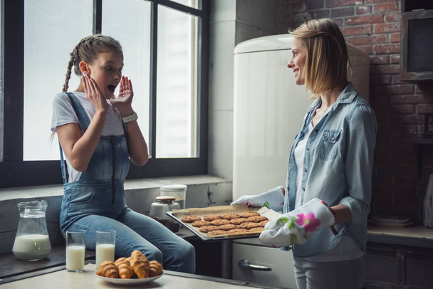 Mom and daughter in kitchen at home, woman is holding a sheet pan with freshly baked cookies, girl is surprised - Fotó, kép