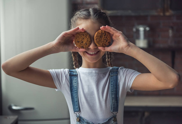 Portrait of little girl holding cookies on her eyes, posing at camera and smiling while standing in kitchen at home - Foto, afbeelding