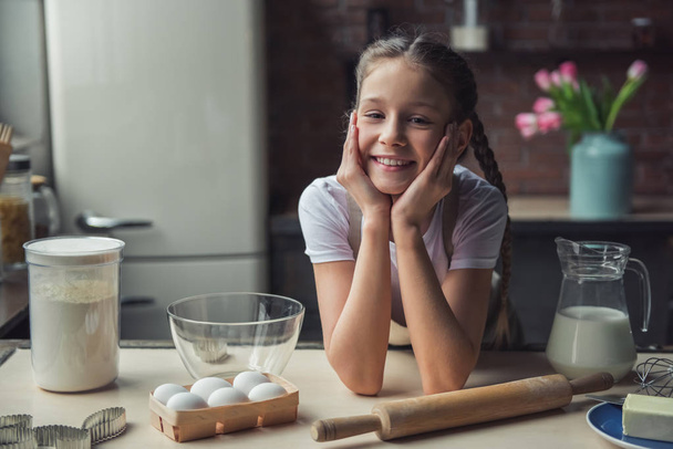 Roztomilá holčička v zástěře je opřena o stůl, při pohledu na fotoaparát a usmíval se při vaření v kuchyni doma - Fotografie, Obrázek