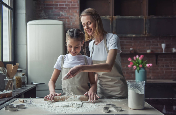 Красивые мама и дочь в фартуках используют скалку и улыбаются, готовя тесто дома
 - Фото, изображение