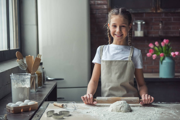 Aranyos kislány kötény látszó-on fényképezőgép, és mosolyogva, miközben egyengető tészta használ a sodrófa, konyha otthon - Fotó, kép