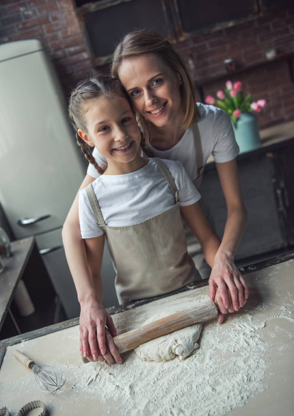 Beautiful mom and daughter in aprons are using rolling pin, looking at camera and smiling while preparing dough at home - Photo, image
