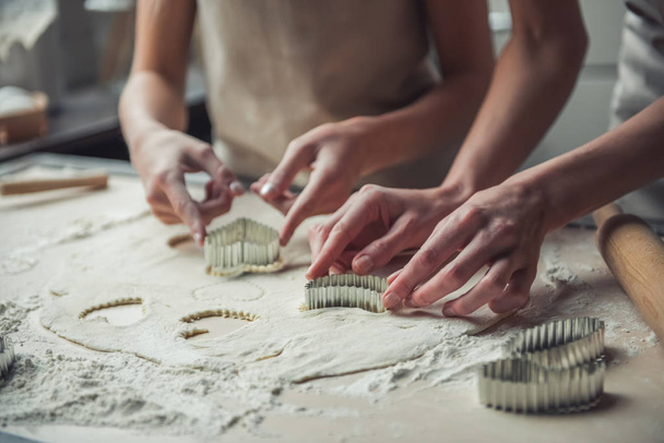 Immagine ritagliata di mamma e figlia in grembiuli utilizzando taglierine di pasta di diverse forme per fare i biscotti
 - Foto, immagini