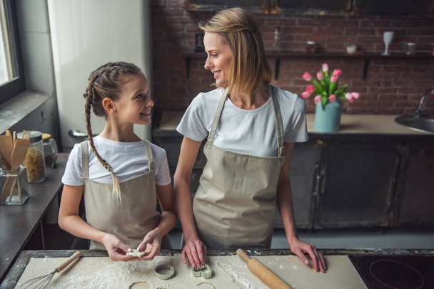 Piękna Mama i córka w fartuchy są patrząc na siebie nawzajem i uśmiechając się podczas używania frezów ciasta do cookies w domu - Zdjęcie, obraz