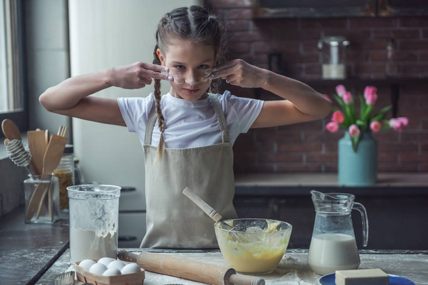 Menina bonito no avental está fazendo linhas em seu rosto com farinha pronta para cozinhar, na cozinha em casa
 - Foto, Imagem