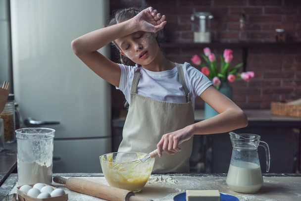 Cute tired little girl in apron is using a whisk and wiping her forehead while preparing dough, in kitchen at home - Foto, Bild