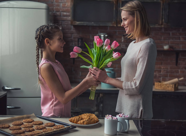 Little girl is giving flowers to her mom, both are smiling while standing in kitchen at home - Photo, Image
