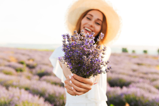 Happy young girl in straw hat holding lavender bouquet at the field - Фото, изображение