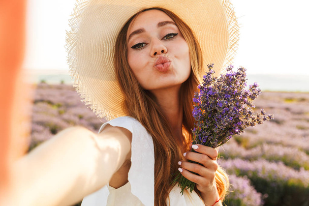 Cheerful young girl in straw hat holding lavender bouquet at the field, taking a selfie - Foto, immagini