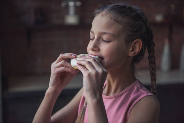 Cute girl is tasting donut with closed eyes while standing in kitchen - Photo, Image
