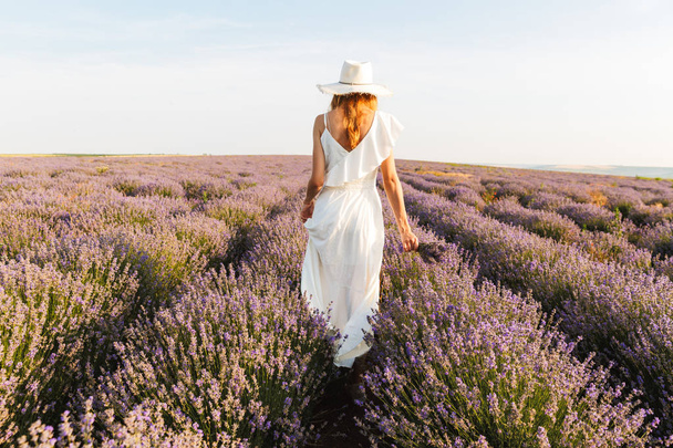 Back view of a pretty young girl in straw hat walking at the lavender field - Photo, Image