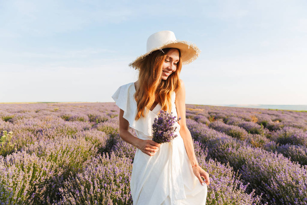 Cheerful young girl in straw hat holding lavender bouquet at the field - Valokuva, kuva