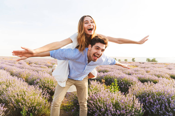 Happy young couple having fun at the lavender field together, piggyback ride - Фото, изображение