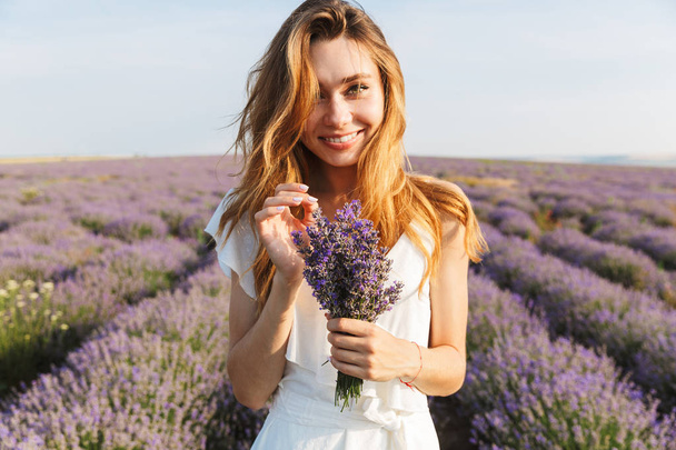 Portrait of cheerful pretty woman in dress holding bunch of flowers while walking outdoor through lavender field in summer - Foto, Bild