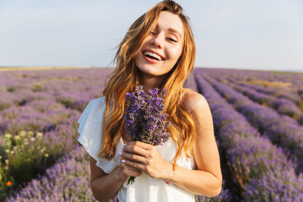 Photo of smiling brunette woman in dress holding bouquet with flowers while walking outdoor through lavender field in summer - Фото, изображение