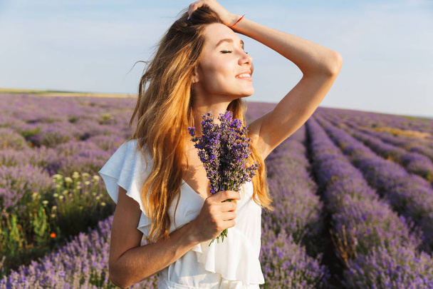 Photo of pretty european woman in dress holding bouquet with flowers while walking outdoor through lavender field in summer - Φωτογραφία, εικόνα