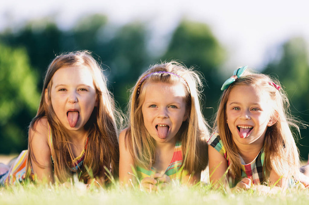 Три маленькие девочки высовывают языки, лежат на траве в парке. Маленькие повстанцы
.  - Фото, изображение