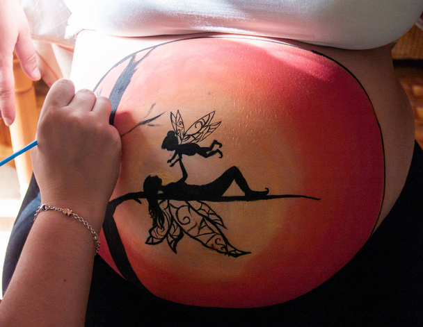Vrouwelijke kunstenaar, schilderij van de buik van een zwangere jongedame met lichaam verf en kwasten - Foto, afbeelding