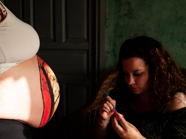 Artiste féminine peignant le ventre d'une jeune femme enceinte avec des peintures corporelles et des pinceaux
 - Photo, image