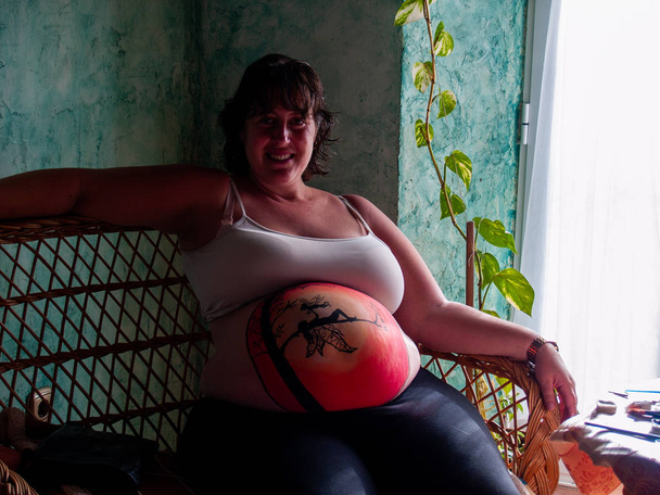 Body Art Jeune femme enceinte souriante avec le ventre peint avec un dessin de fée, assise sur un canapé
 - Photo, image