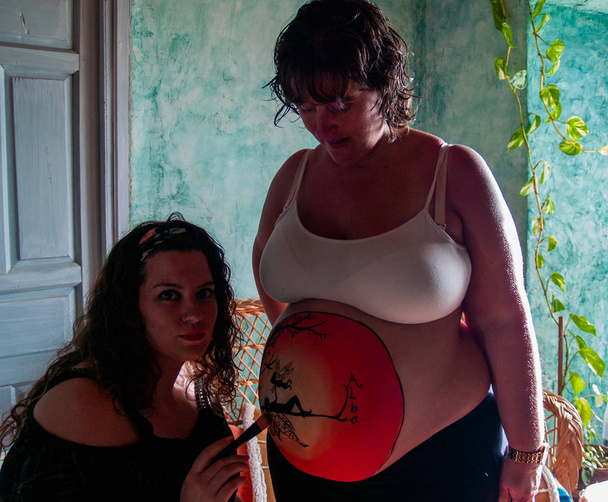 Artiste féminine peignant le ventre d'une jeune femme enceinte avec des peintures corporelles et des pinceaux
 - Photo, image