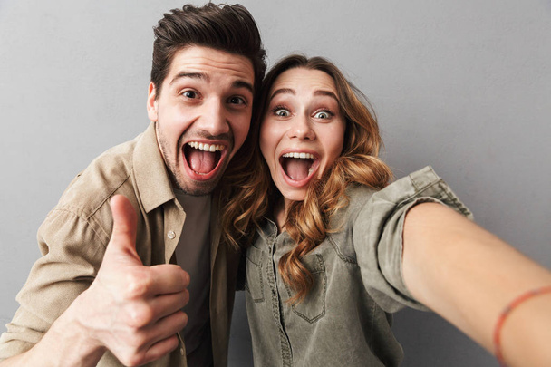 Portrait of a happy young couple hugging while taking a selfie and showing thumbs up isolated over gray background - Fotografie, Obrázek