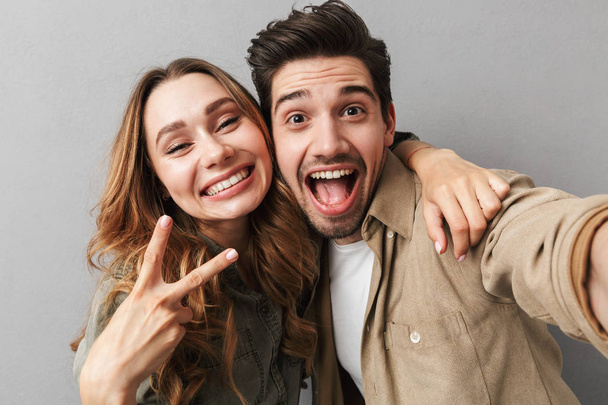 Portrait of a cheeful young couple hugging while taking a selfie and showing peace gesture isolated over gray background - Photo, Image
