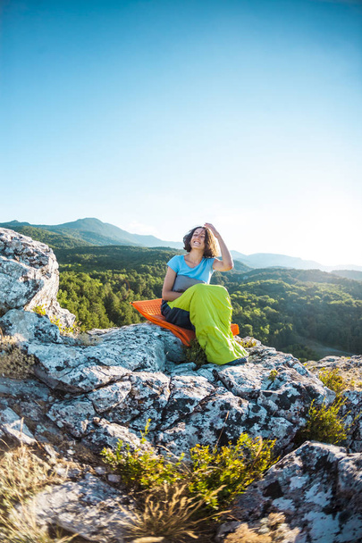 Žena sedí ve spacáku na vrcholu hory. Usmívající se dívka odpočívá po stoupání. Brunetka se nachází na sebe hustí tácek, na pozadí pohoří a zapadajícího slunce. - Fotografie, Obrázek
