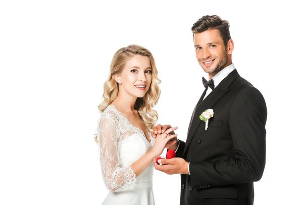 happy groom putting on wedding ring on brides finger and looking at camera isolated on white - Photo, Image