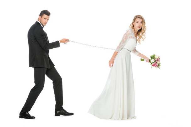 side view of young groom with chain and leashed shocked bride isolated on white - Φωτογραφία, εικόνα
