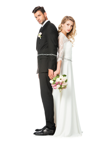 serious young newlyweds tied with chain back to back and looking at camera isolated on white - Foto, Bild