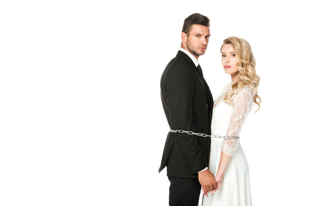 serious young newlyweds tied with chain and looking at camera isolated on white - Фото, изображение