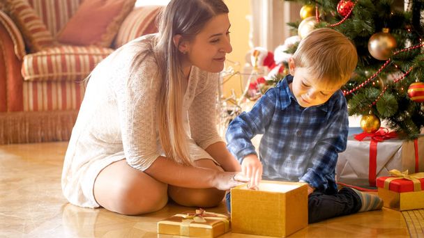 Little toddler boy sitting with mother under Christmas tree and opens magical glowing gift box - Foto, Bild