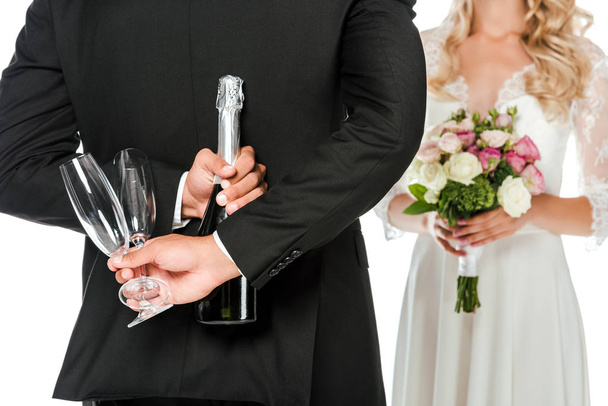 cropped shot of groom holding champagne bottle and glasses behind back while bride standing in front of him isolated on white - Photo, Image