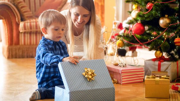 Cute toddler boy looking inside of CHristmas gift box with young mother - Photo, Image
