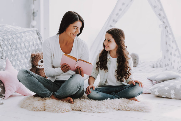 Young Mother Reading Book to Little Daughter. Beautiful Smiling Woman Sitting on Carpet Reading to Cute Adorable Daughter. Mom reads to Child at Home. Family and Motherhood Concept - Foto, Imagen