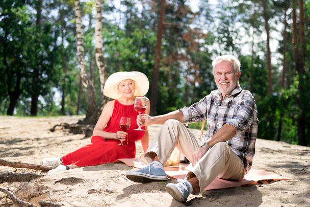 Couple of beaming elderly wife and husband sitting on sand having picnic - Photo, Image